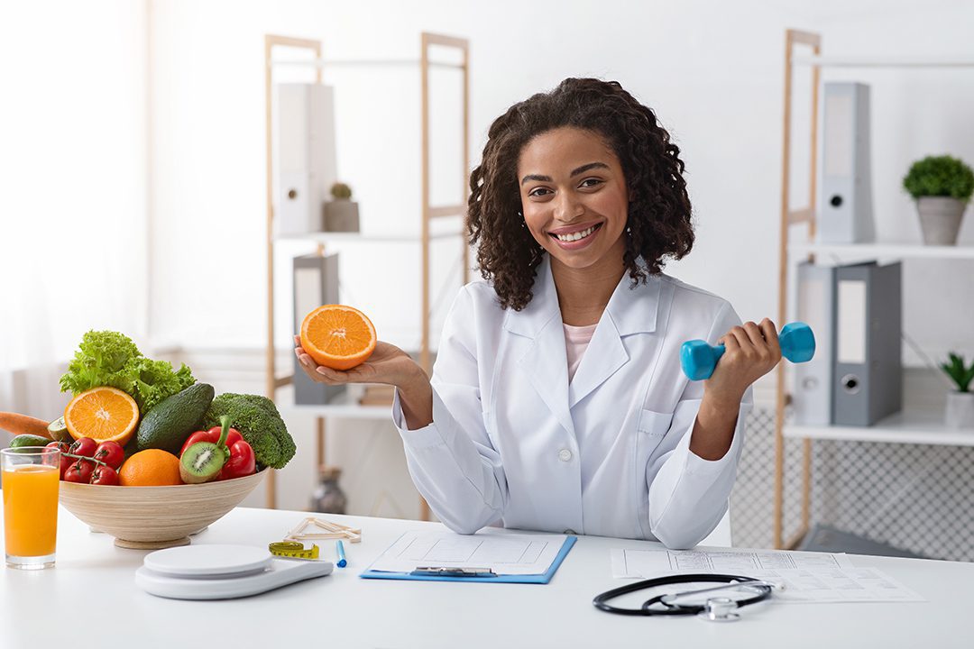 Young cheerful afro woman dietologist holding orange half and dumbbell in her hands, recommending healthy lifestyle